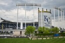 A large baseball stadium sits behind a manicured lawn. A large sign reads "KC 1985, 2015 World Champions."