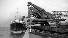 A Great Lakes freighter is pulled up against a bulkhead near four massive mechanical iron ore unloaders that look like giant metal elephants or praying mantises. 