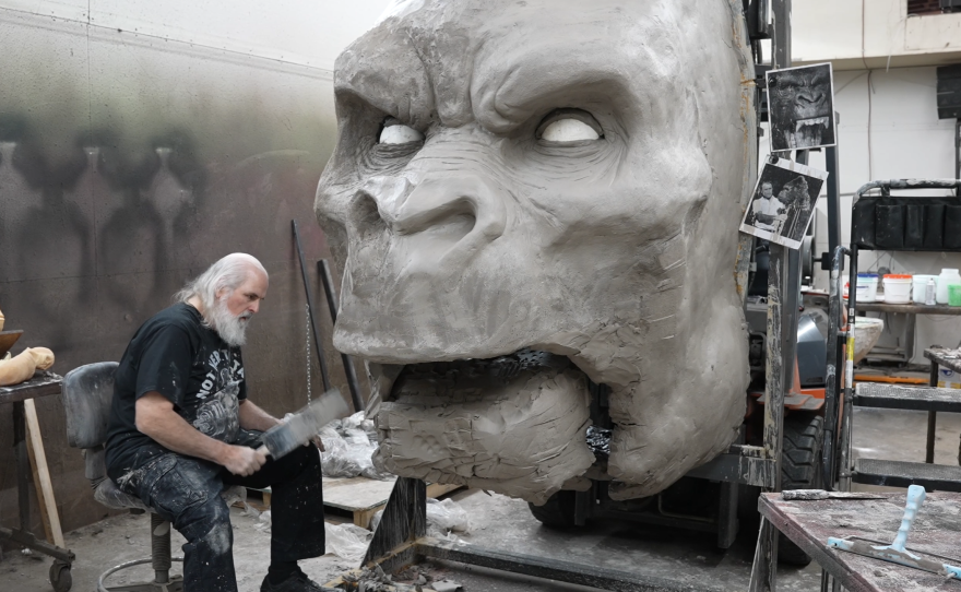 Ed Edmunds sits in front of a massive gray sculpted monster head with white eyes