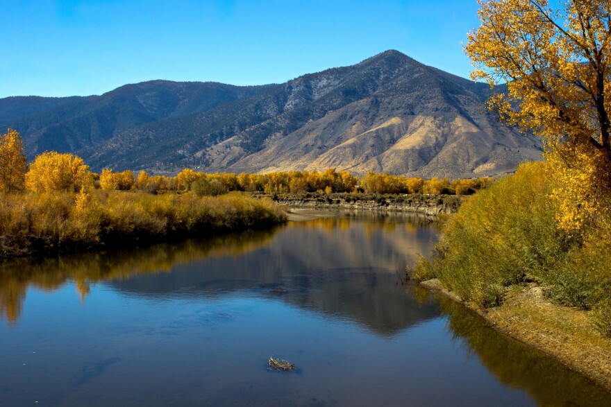 Fall colors reflect in a river on a clear blue-sky day. A mountain range is in the backdrop. 