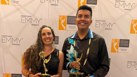 Two dressed-up people hold gold statues and smile at the camera, in front of a backdrop with the words “Heartland Emmy Awards” repeated across the background.