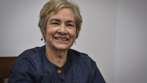 A woman sits looking at the camera and smiling. She is sitting in a wooden chair in a room with a white wall.