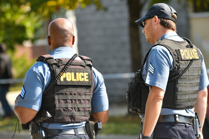 Two police officers wearing blue shirts and black vests with "Police" printed on the back stand near a crime scene.