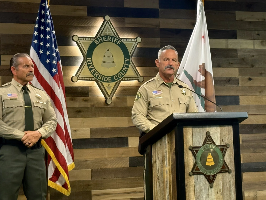 A man in a sheriff's uniform stands behind a podium next to another man in the same uniform. 