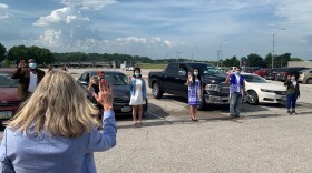 Four people stand in front of their cars holding up their right hand. A woman stands in front of the camera so you see the back of her head. She also holds her right hand up.