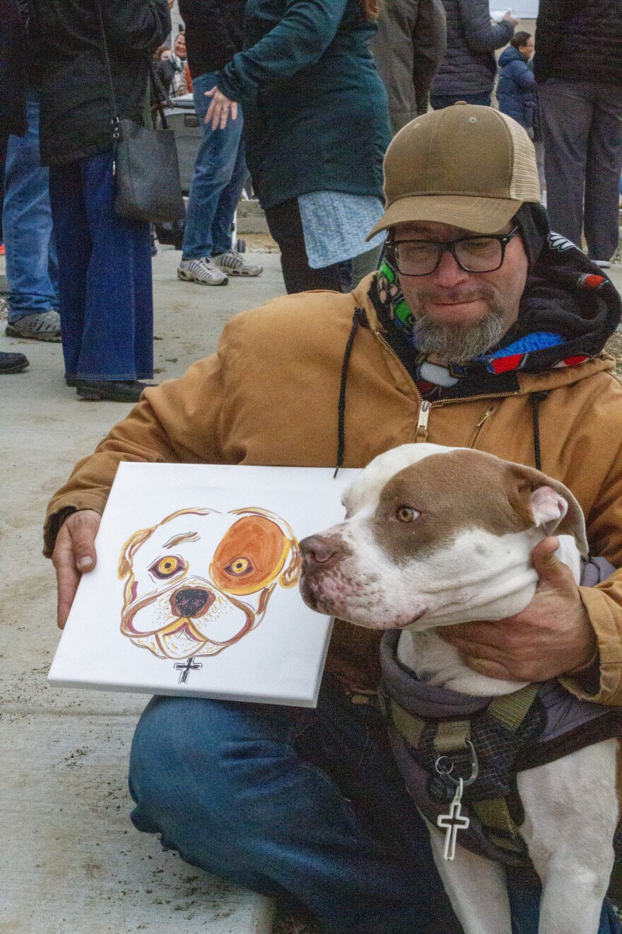 A man crouches with his dog, holding a hand painted portrait of the pup.