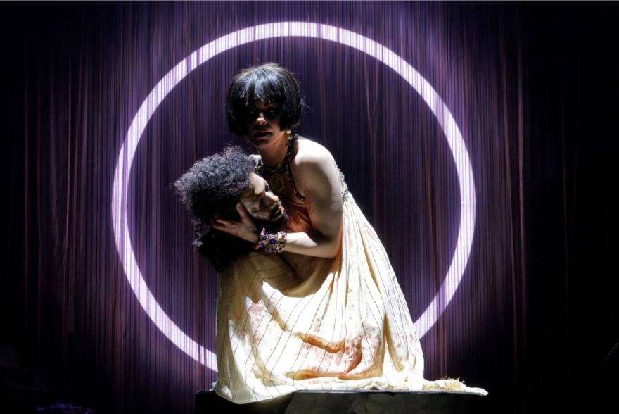 A female holds a male in her arms, with blood on her white dress.