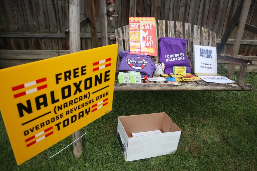 a bright yellow sign reads "free naloxone" next to a bench that holds supplies