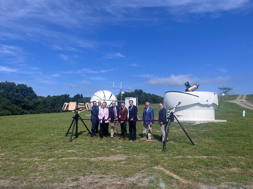 8 men and one woman stand in front of two large telescopes, hosting a ribbon that is being cut with a pair of scissors.  