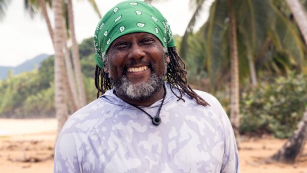 African American man with greyish beard and brown dreads wears a bandana on beach smiles into camera 