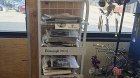 An in-store newsstand with stacks of newspapers placed inside of a window.