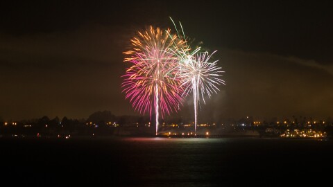 Circular pink, orange and light pink fireworks overlap with some flashes of green on a dark night with small lights illuminating a black background. 