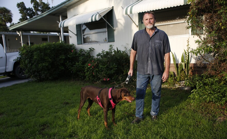 An elderly man wearing a shirt and jeans stands outside his home with his dog on a leash. 