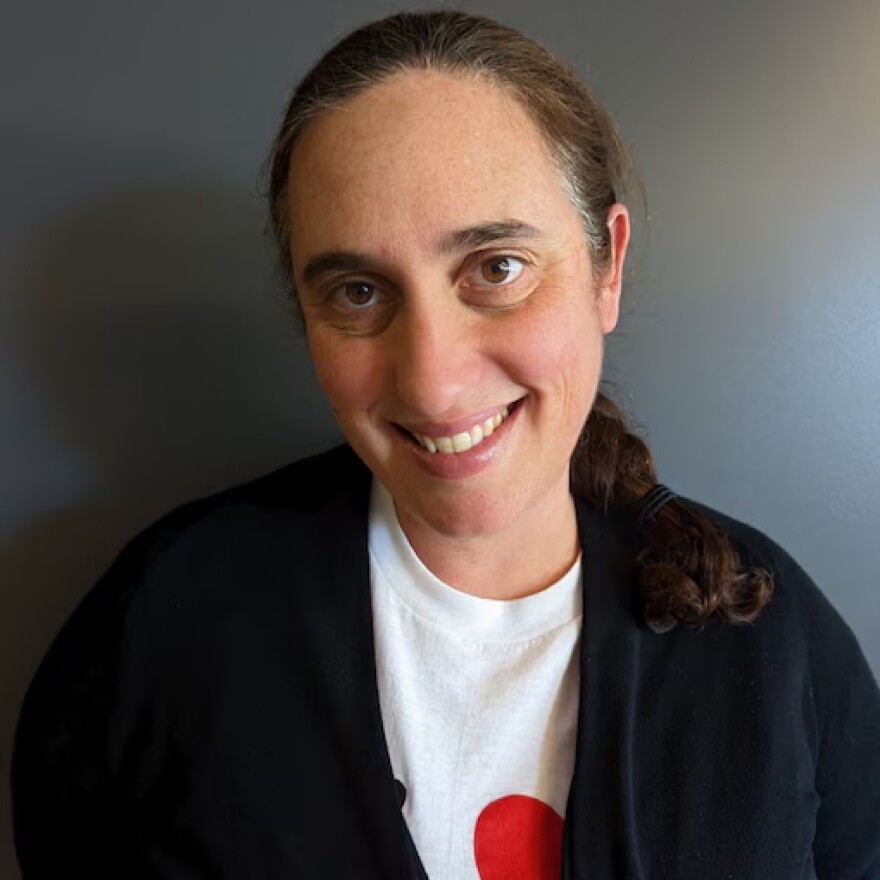A woman is smiling at the camera. She has long brown hair and wears a white shirt and black cardigan. There is a gray wall behind her. 