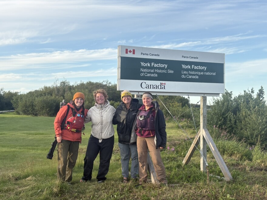 Four women stand beneath a sign that reads "York Factory: National Historic Site of Canada." Green grass and small bushes surround them. They are wearing wind breakers and two are wearing knitted hats. 