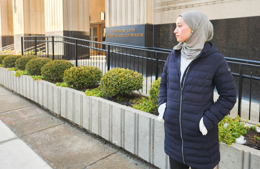A young woman in a gray hijab and navy coat stands in front of the Theodore Levin United States Courthouse in Detroit. Behind her is a railing and decorative shrugs. She is looking to the left of frame. 