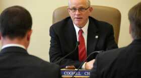 A bald man in a black suit with a white tie talks to two other men (with their backs turned) at a desk.