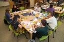 Participants sit around tables covered with paper, photographs, sticky notes, scissors and markers during a creative workshop in a library or classroom setting. Several groups work on hands-on projects while computers line the back wall.