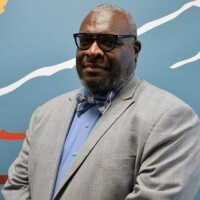 A bust shot of Sam Radford, wearing a light blue suit, shirt and bow tie, in front of a blue, white and orange striped background.
