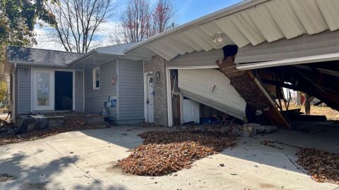 An abandoned home. It's grey with beige bricks and white trim. The garage to the right is destroyed.