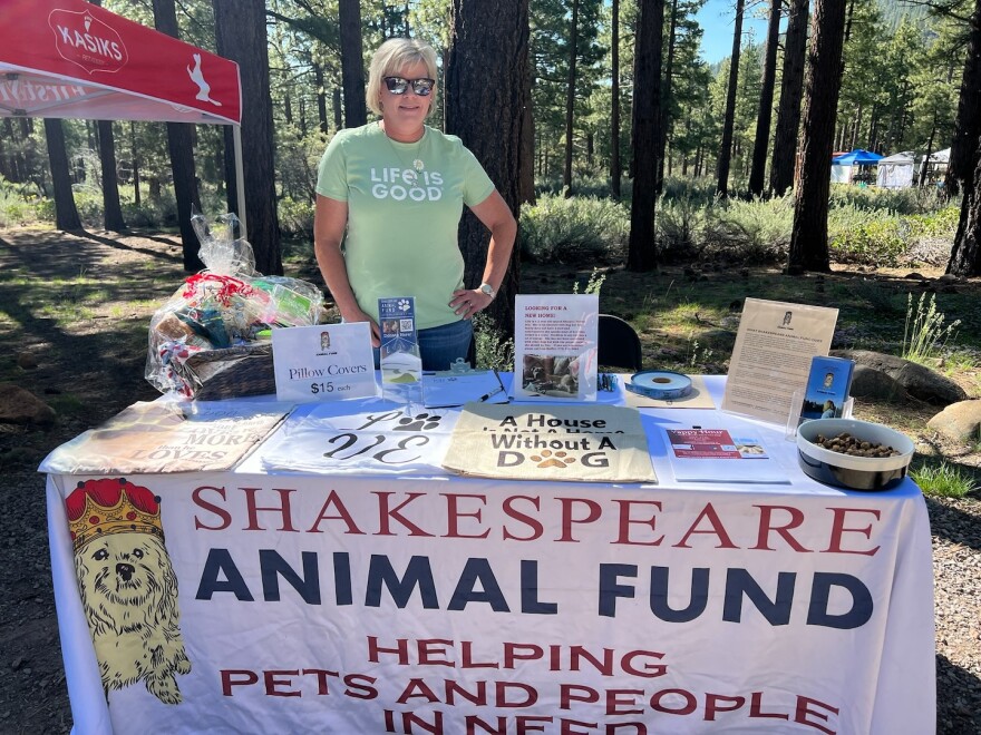 A table set up outside. There is a tablecloth with animal-related posters and merchandise set on the table. A woman is standing behind the table while smiling and looking toward the camera.