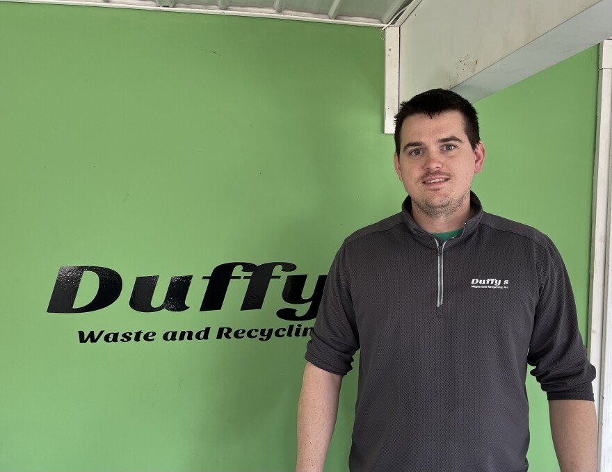 A man stands in front of a green wall with the words "Duffy's Waste and Recycling" printed on it.