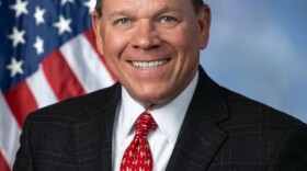 A man, Rep. Sam Graves of Missouri's 6th District, is dressed in a black suit and red tie, smiling, seated for a portrait in front of a draped American flag backdrop.