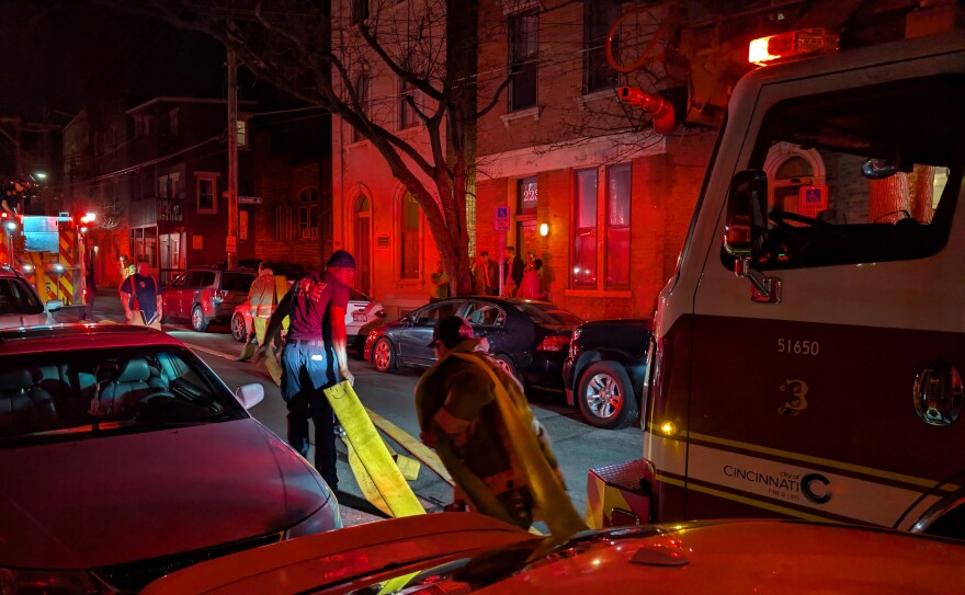 Firefighters drag hoses in a dark street lit up by fire truck flashing lights.