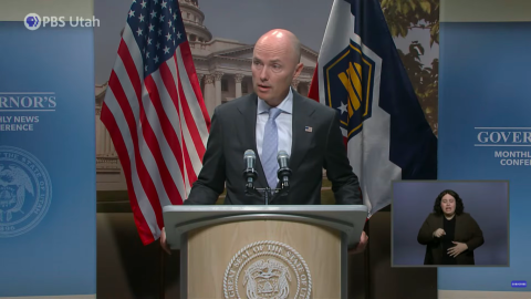 Utah Governor Spencer Cox stands behind a podium backed by the American and Utah state flags. 