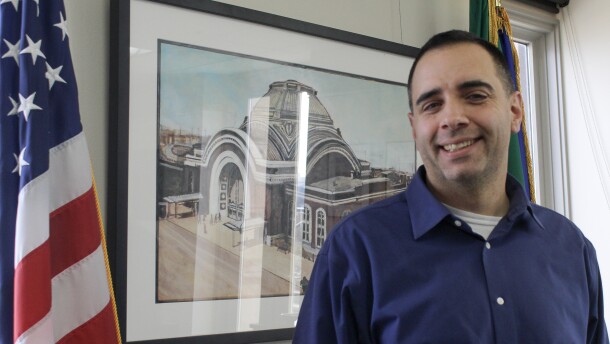 Tacoma Mayor Anders Ibsen stands in front of a painting and alongside the U.S. and Washington flags.