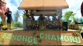 A float with a thatched roof and floral decorations displays the words “Fanohge CHamoru” during Guam’s 81st Liberation Day parade. People sit inside the float, and a man in black stands at the front, calling out to the crowd.