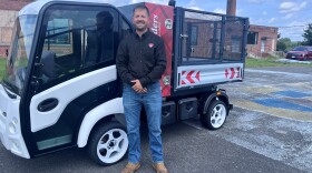 A man wearing a black shirt and jeans stands in front of a small white truck.