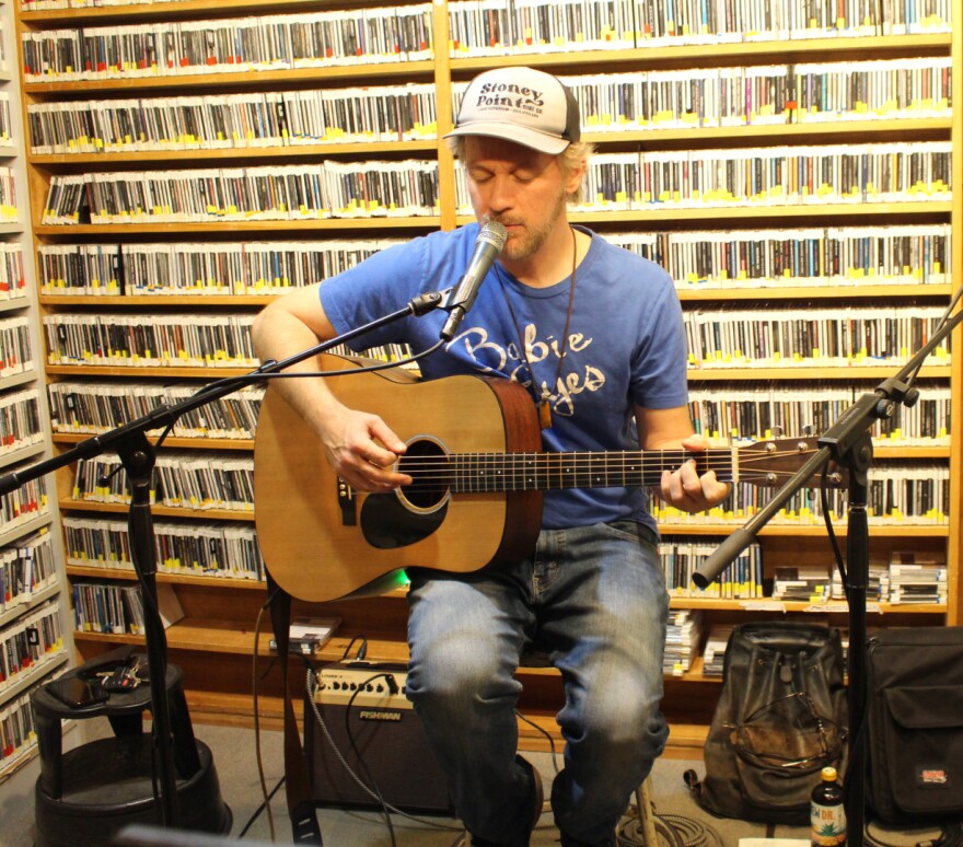 A seated man plays guitar and sings into a microphone in a radio studio. Behind him and to the left are shelves full of CDs lining the walls.