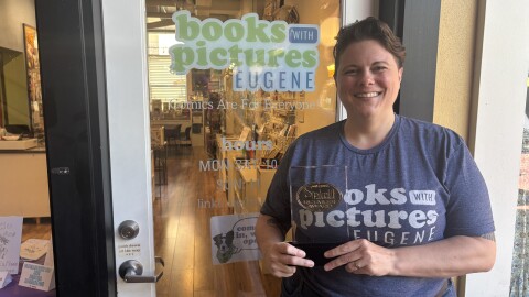 Andréa Gilroy, the shop owner, standing in front of 'Books with Pictures', holding their newly won Will Eisner Spirit of Retail Award in Eugene, Ore. 7/27/2026