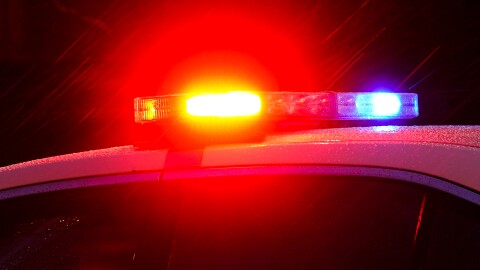 Shown are the red and blue flashing lights of a police vehicle, zoomed in to show only the lights and the top half of the police cruiser. Rain is trickling down through the frame, illuminated by the flashing lights. 