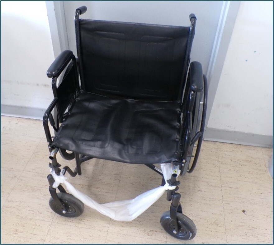 A white plastic bag is tied across the bottom of a black wheelchair at Women's Huron Valley Correctional Facility, the state's only women's prison where the footrest should be. The wheelchair is on a brown tiled floor against a white wall.