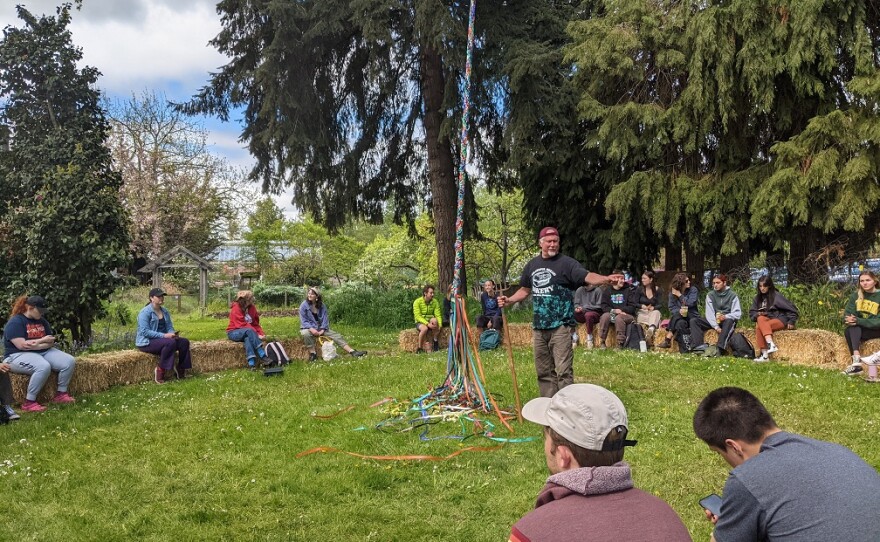 A man with a pitchfork stands in  clearing with students sitting on hay bales in a circle around him. A May pole is next to him. Behind him are tall cedar trees. 