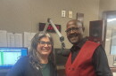 On the left, Mona Seghatoleslami smiling wearing a green dress and black jacket. On the right, viola professor George Taylor, also smiling, wearing a black shirt with a red vest. They are standing in a studio in front of a microphone and a computer.