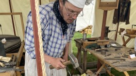 A man in traditional clothing is bent over a piece of metal, holding a hammer. He is inside a tent filled with old-fashioned tools.