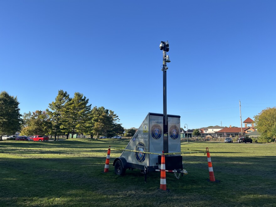 Camera on the top of a long pole, on a mobile trailer with solar panels on the bottom. Pictured in the middle of a grassy field. 
