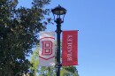 A black lamp pole, featuring a white banner with the Bradley University logo on the left and a red banner with "Bradley" spelled out on the right, stands beneath a bright blue sky in front of a row of trees on the campus.  