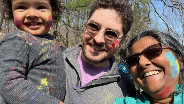 A man and woman pose with a child. All three have paint splattered on parts of their face.