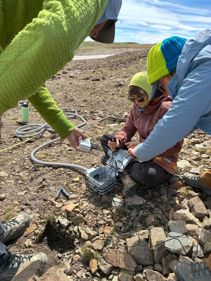 Three people in cold-weather gear on rocky ground. One of the scientists is sitting with the tool they're using, which is an open box with a thick cable running to somewhere not visible and a series of smaller wires coming out of the box. Another scientist is standing nearby holding the little monitor those wires are connected to, and another is holding onto some of the wires.