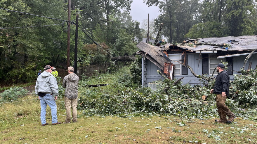 Three men stand among tree brush that surrounds a house. Part of the roof is caved in from a falling tree.