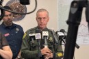 A sheriff standing up with several microphones pointed toward him. He is looking away from the camera.