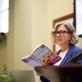 A woman with short blonde hair and black glasses wears a blue blazer and reads a book titled "Smog and Sunshine"