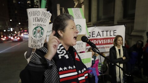 A woman with short gray hair wearing a "Washington Democrats" scarf speaks into a microphone on a sidewalk where protesters hold signs, one of which is shaped like a Starbucks cup and read "Third Party Howards Equals Trump Again!"