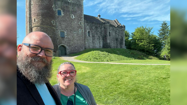 Two people posing for a picture in front of grey castle with a lot of green grass surrounding it.