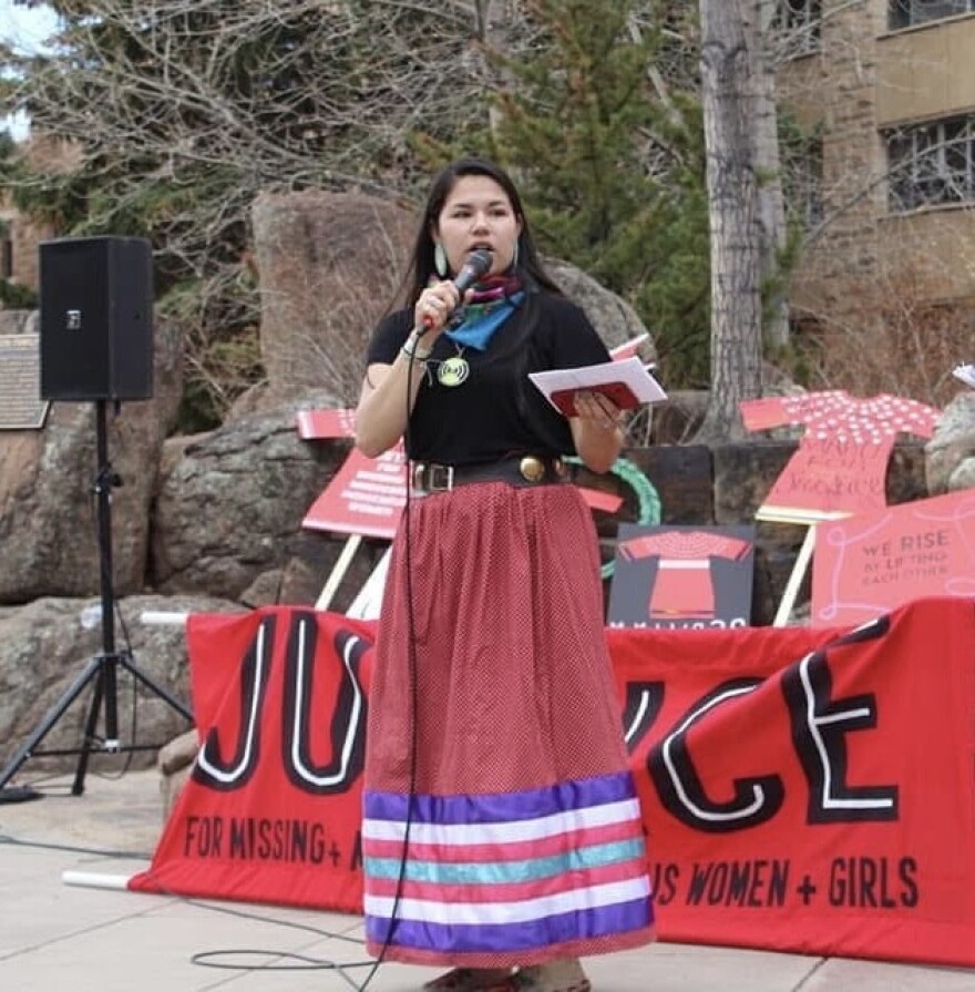 A young woman wearing a black shirt and ribbon skirt speaks into a microphone outside at a college campus. She stands in front of signs with red dresses and a red banner with the word “Justice” on it.