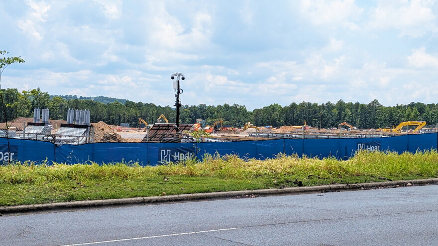 A construction site is visible above a blue privacy fence. Concrete pilings, yellow construction machines and a security camera are visible.